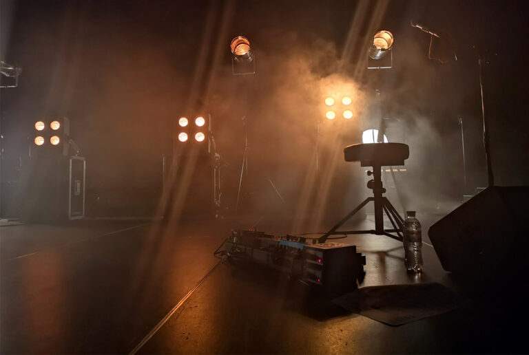 Un escenario iluminado por luces cálidas y envuelto por equipo técnico musical evocando la magia de la música y conciertos en vivo.